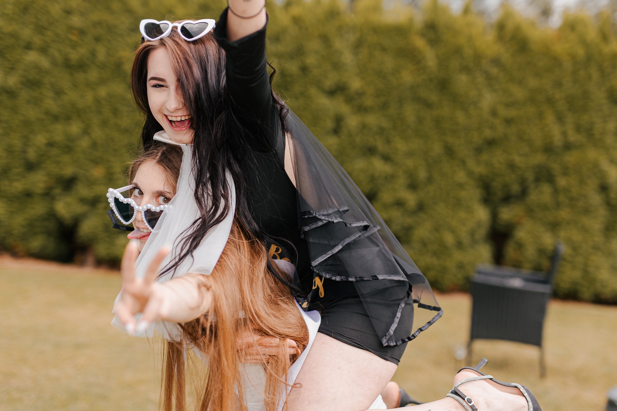 Dwie uśmiechnięte kobiety w okularach w kształcie serc, jedna na barana drugiej, pokazują znak zwycięstwa na tle zieleni ogrodu. Radosna atmosfera.