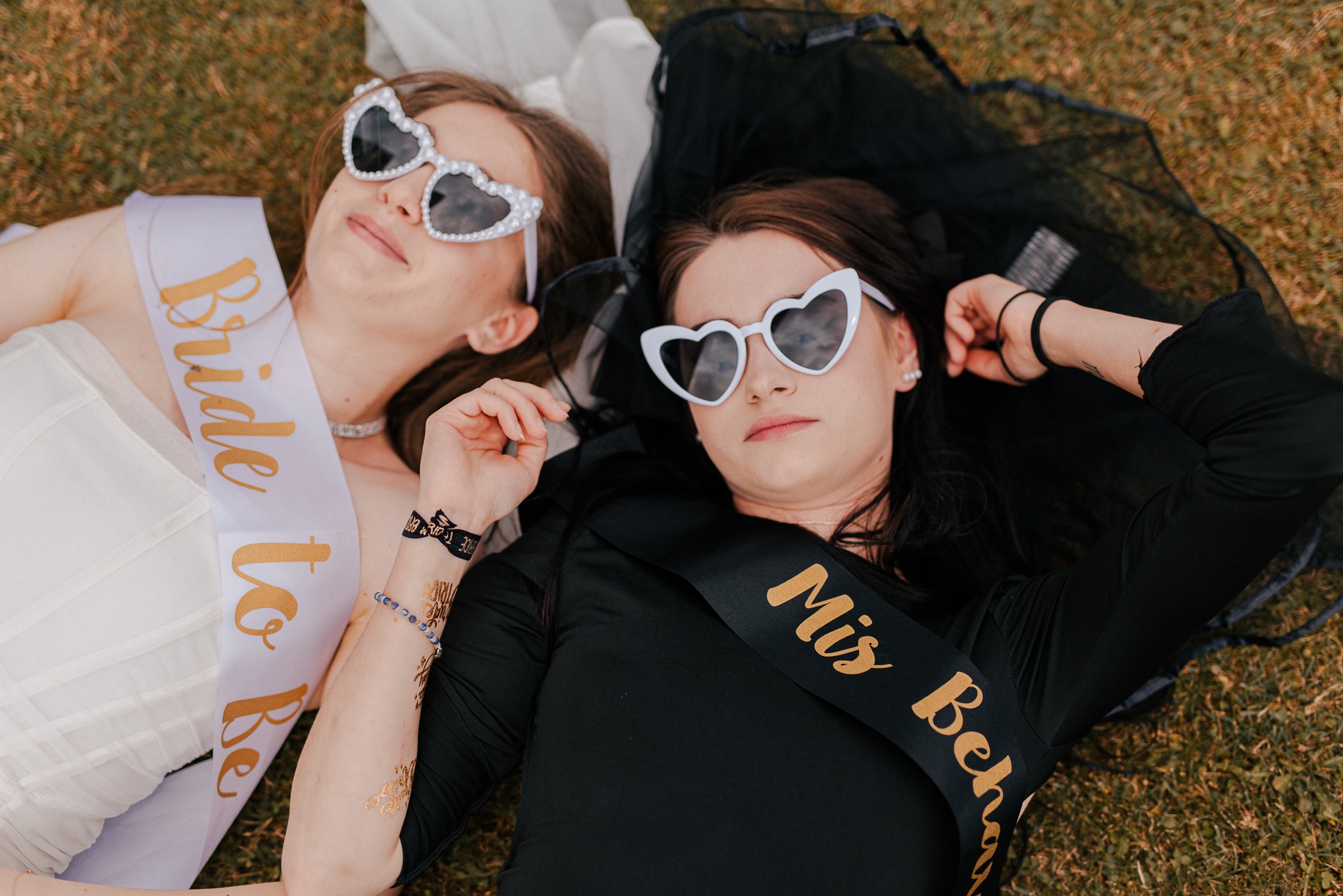 Dwie kobiety w okularach w kształcie serc leżą na trawie. Jedna z szarfą 'Bride to Be', druga 'Mis Behaved'. Uśmiechnięte, w stylizacjach na wieczór panieński.