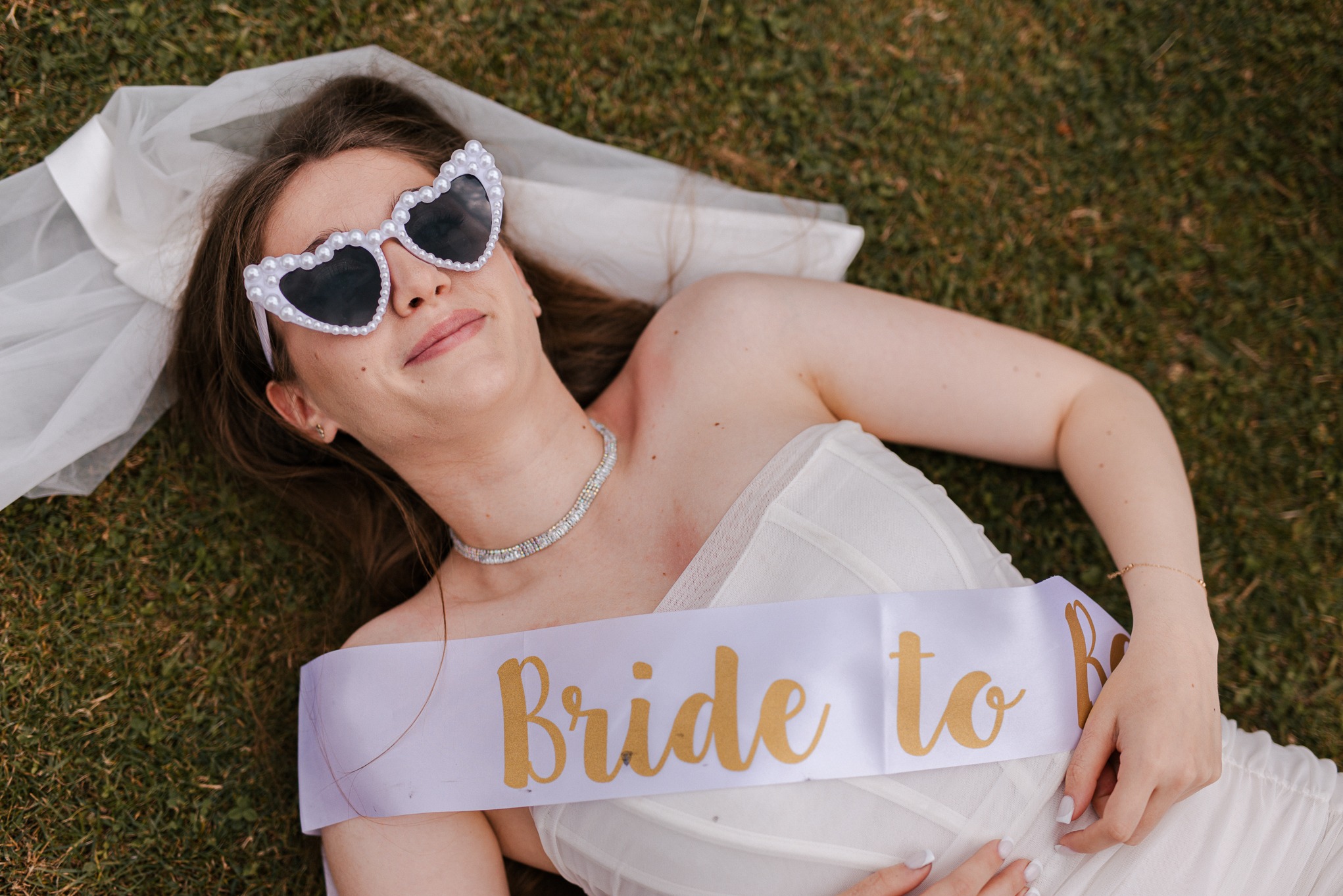 Panna młoda leży na trawie w okularach w kształcie serc i z szarfą 'Bride to Be'. Uśmiechnięta kobieta w bieli, z welonem, w słoneczny dzień. Sesja narzeczeńska w plenerze.