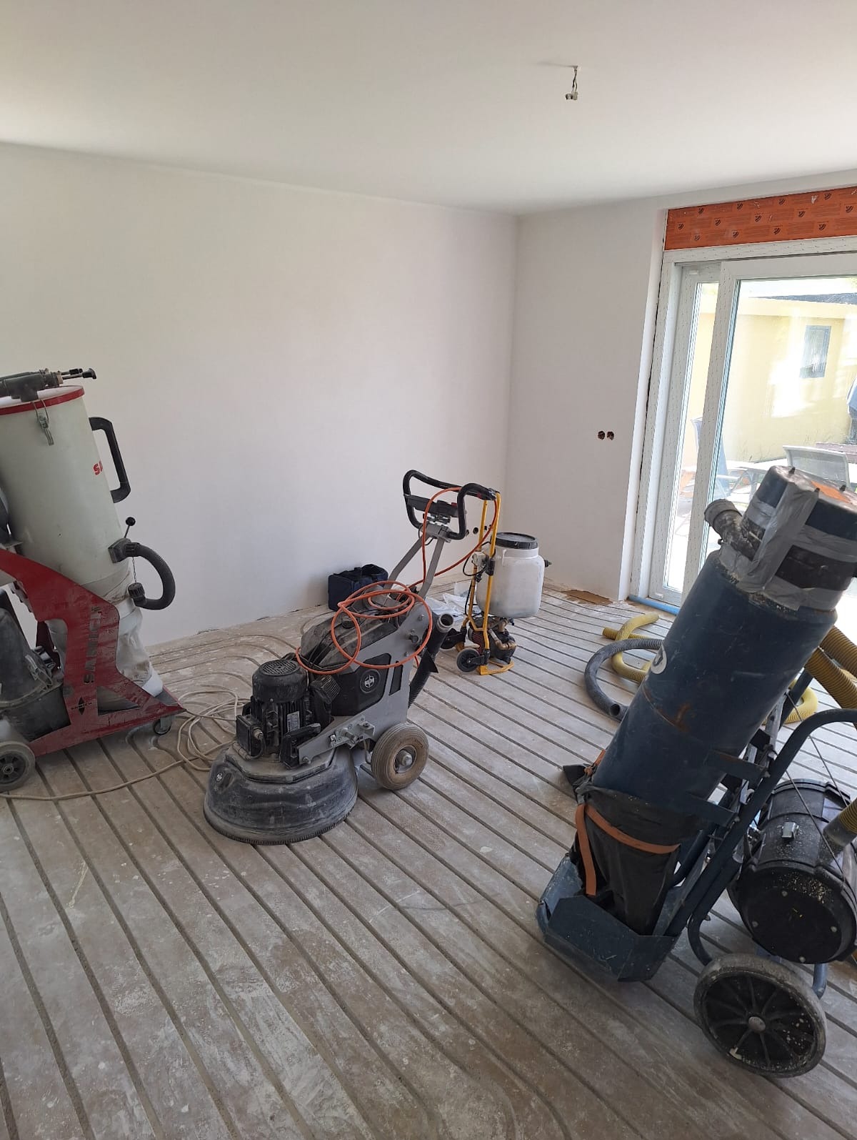 Przygotowanie do wylewki: rury ogrzewania podłogowego ułożone równolegle, widoczne maszyny do wylewania posadzki i odkurzacz przemysłowy. Jasne wnętrze.