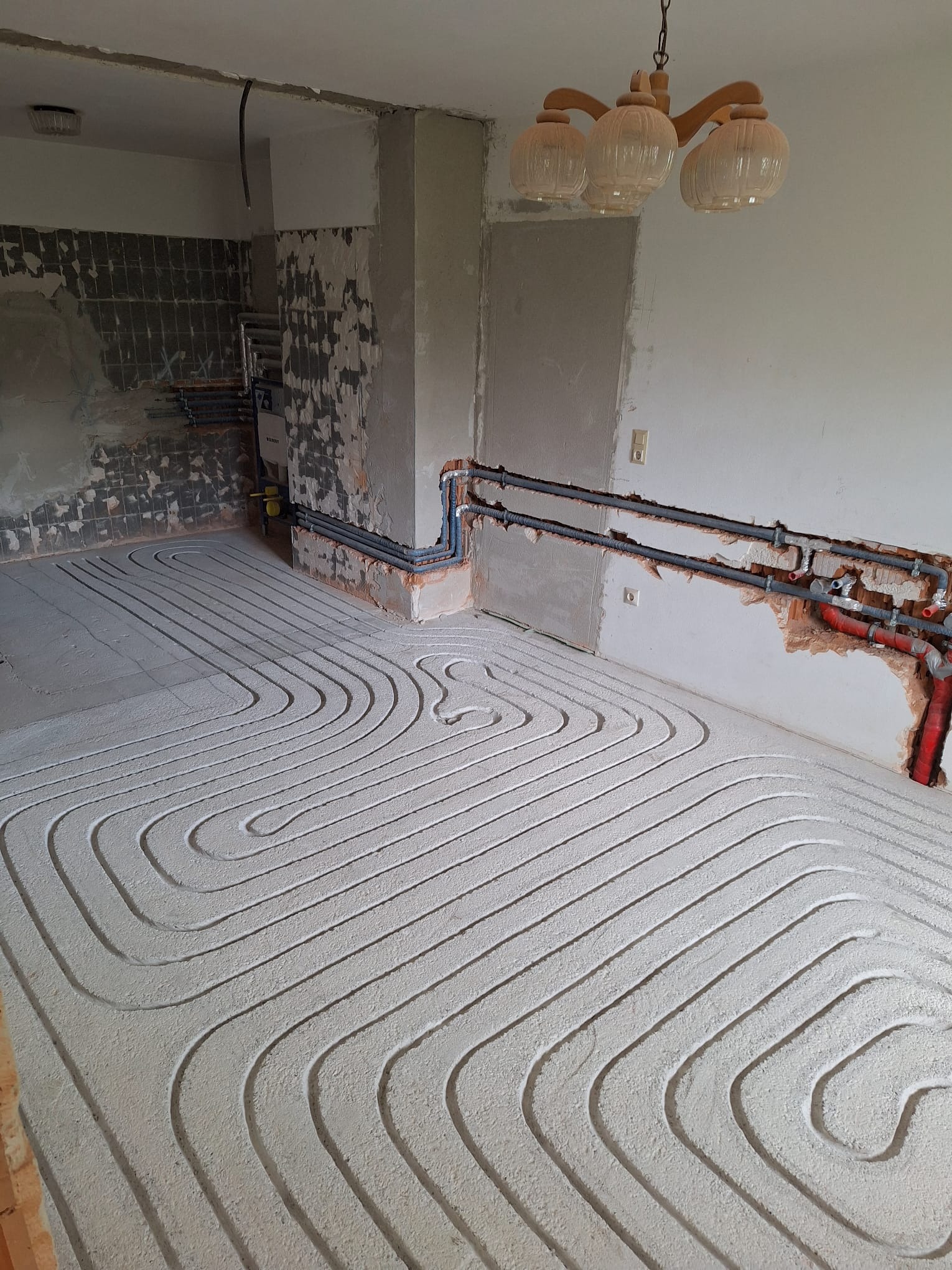 Białe rury ogrzewania podłogowego ułożone spiralnie na podłożu w surowym wnętrzu. Widoczne wystające rury instalacji grzewczej na ścianach.
