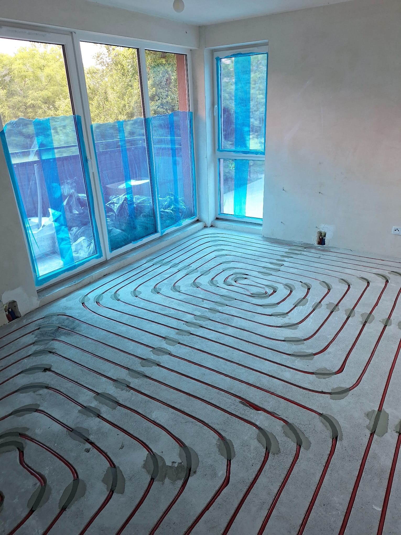 Czerwone rury ogrzewania podłogowego ułożone spiralnie na szarym betonie w jasnym pokoju z oknami zabezpieczonymi folią. Widoczne szare mocowania rur do podłoża.