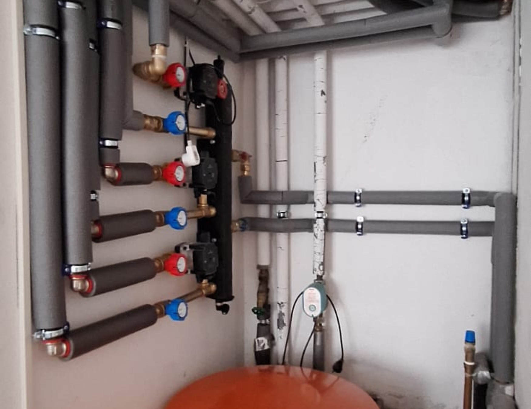 Kompletna instalacja grzewcza z rurami w izolacji, zaworami z kolorowymi pokrętłami i pompami obiegowymi zamontowana w pomieszczeniu technicznym.