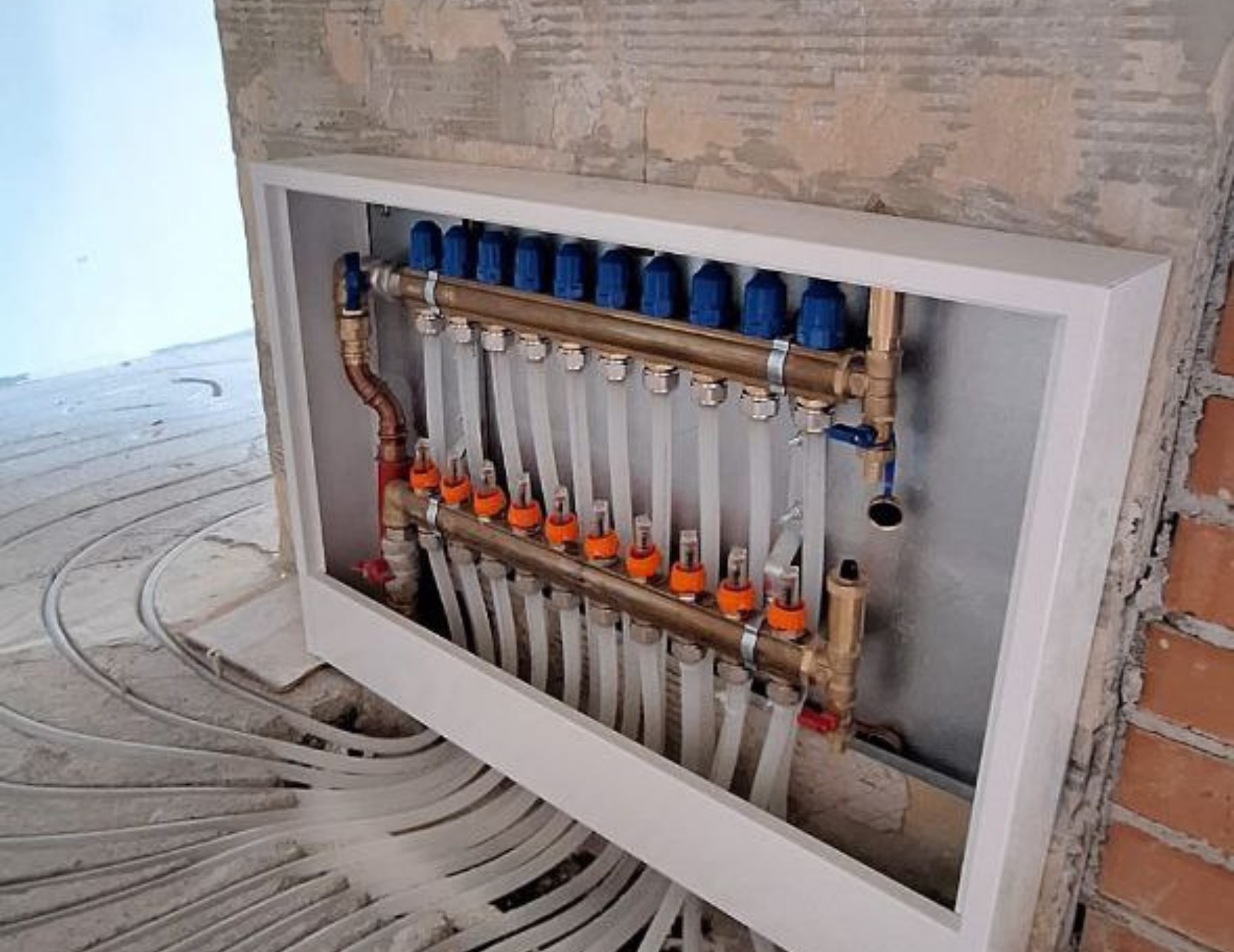 Rozdzielacz ogrzewania podłogowego w białej obudowie, z widocznymi rurami w podłodze. Surowe ściany w tle wskazują na etap instalacji.