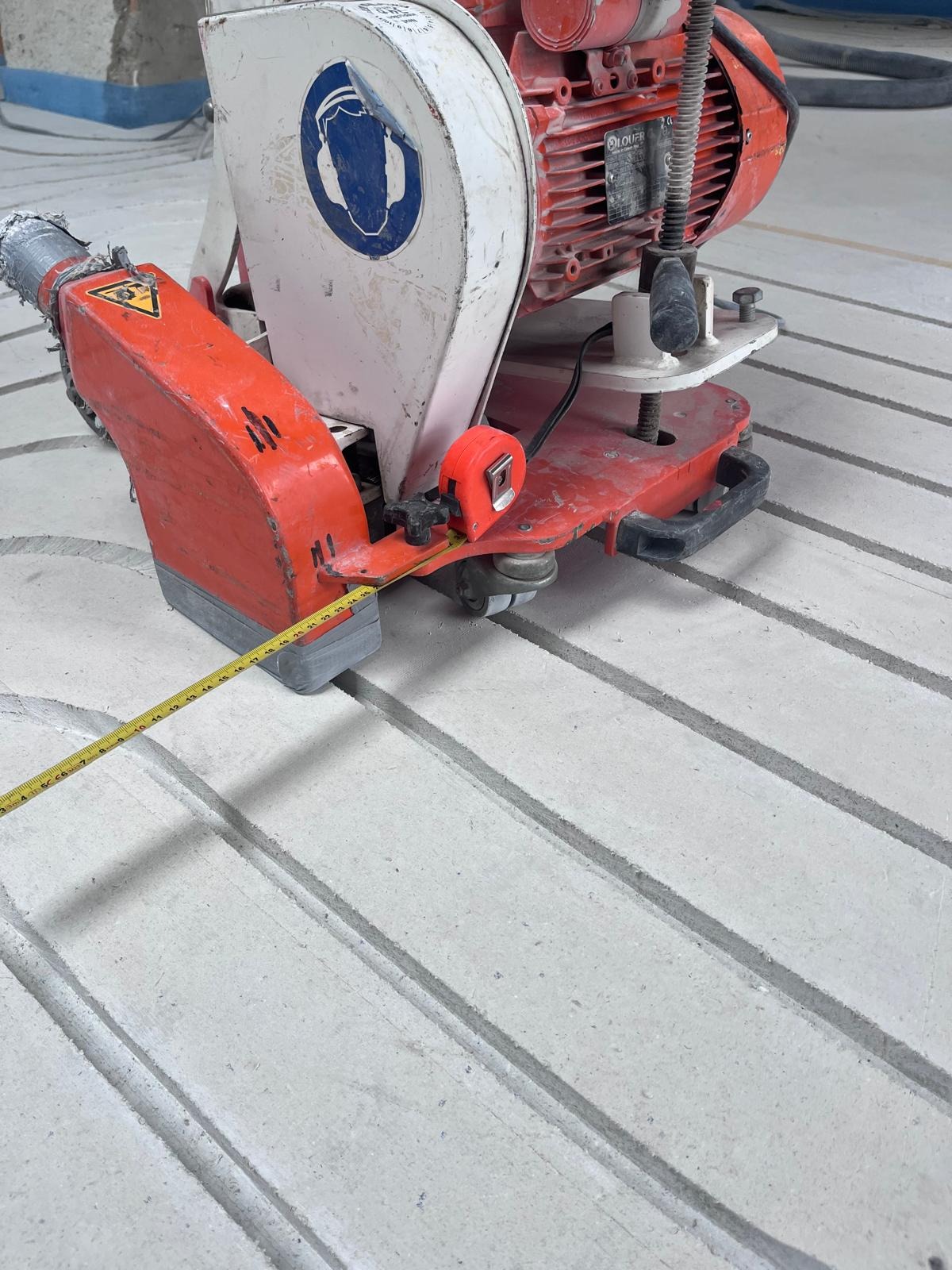 Frezarka do betonu z taśmą mierniczą na szarej, wyfrezowanej podłodze. Widoczne równoległe rowki przygotowane pod instalację ogrzewania podłogowego.