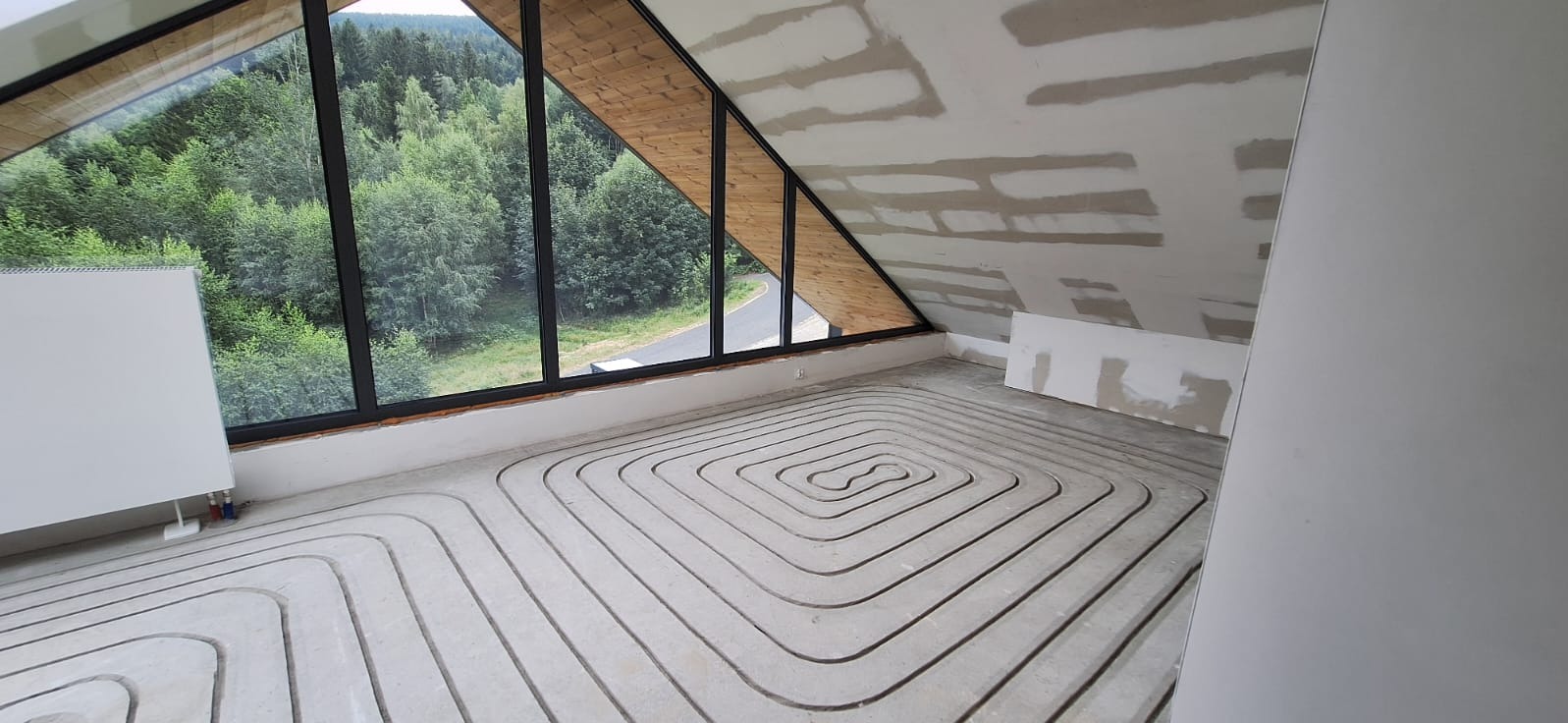 Instalacja ogrzewania podłogowego w surowym wnętrzu poddasza z oknami na las. Widoczne rury w kształcie meandra zalane w betonie, grzejnik i przygotowane płyty gipsowe.