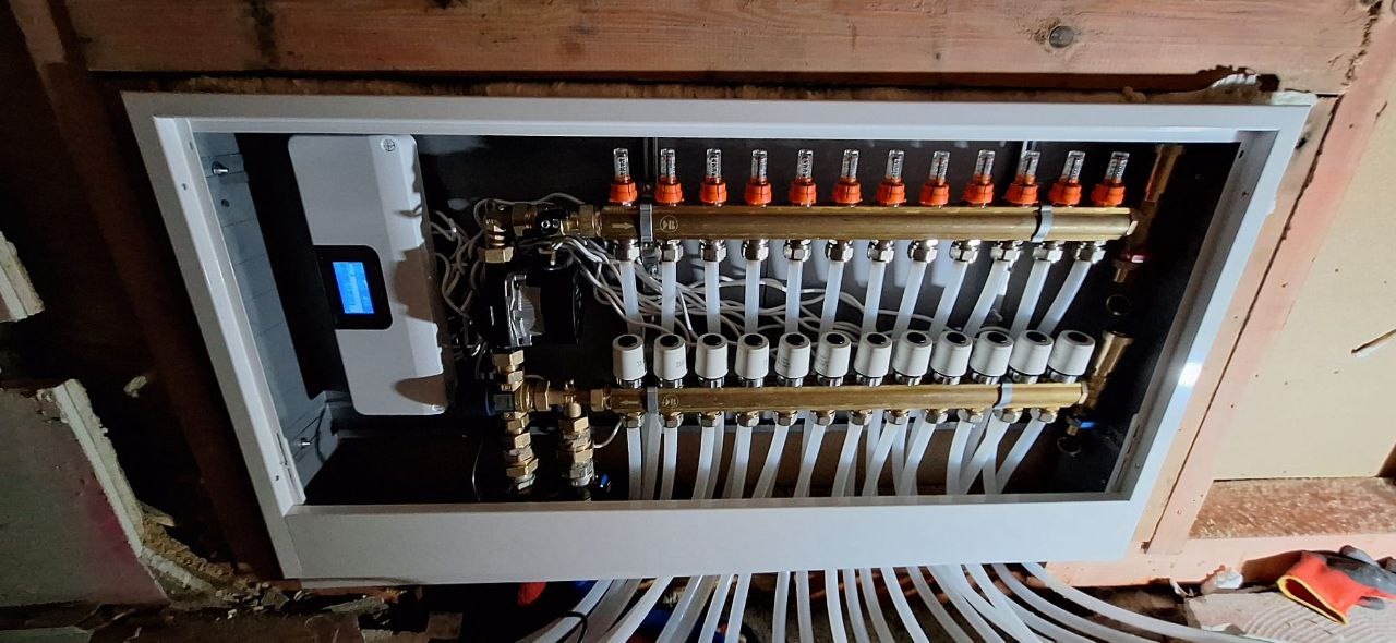 Rozdzielacz ogrzewania podłogowego w białej obudowie, widoczne rury i elementy instalacji, zamontowany na drewnianej ścianie w trakcie prac wykończeniowych.