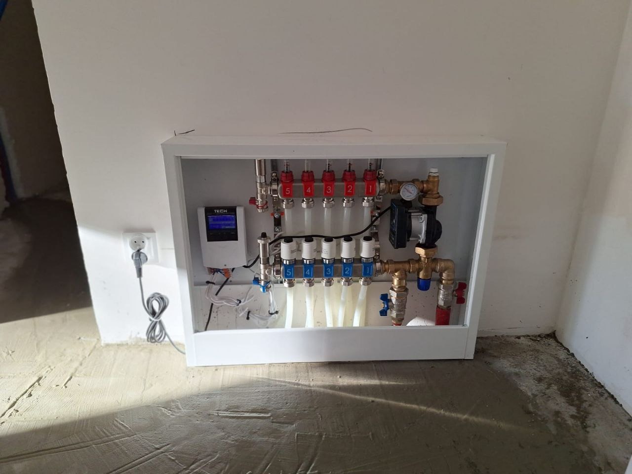 Rozdzielacz ogrzewania podłogowego w białej obudowie, widoczne liczne zawory, rury i manometr. Szare przewody elektryczne i wtyczka w gniazdku.