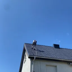 Pracownik w uprzęży na dachu domu z ciemną dachówką, pod błękitnym niebem. Widoczna drabina i fragment białej ściany budynku. Ujęcie z dołu, w słoneczny dzień.