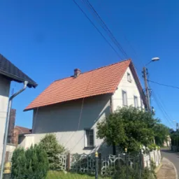 Dwupiętrowy dom z czerwonym dachem i jasną elewacją w słoneczny dzień. Widoczne okna, komin, drzewa i ogrodzenie. Na pierwszym planie trawa i ulica.