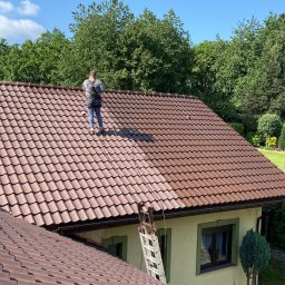 Patryk Mackiewicz - Pracownik z zabezpieczeniami na dachu domu z brązową dachówką, widoczna drabina i częściowo umyty fragment dachu, błękitne niebo. Sezonowe mycie dachu.