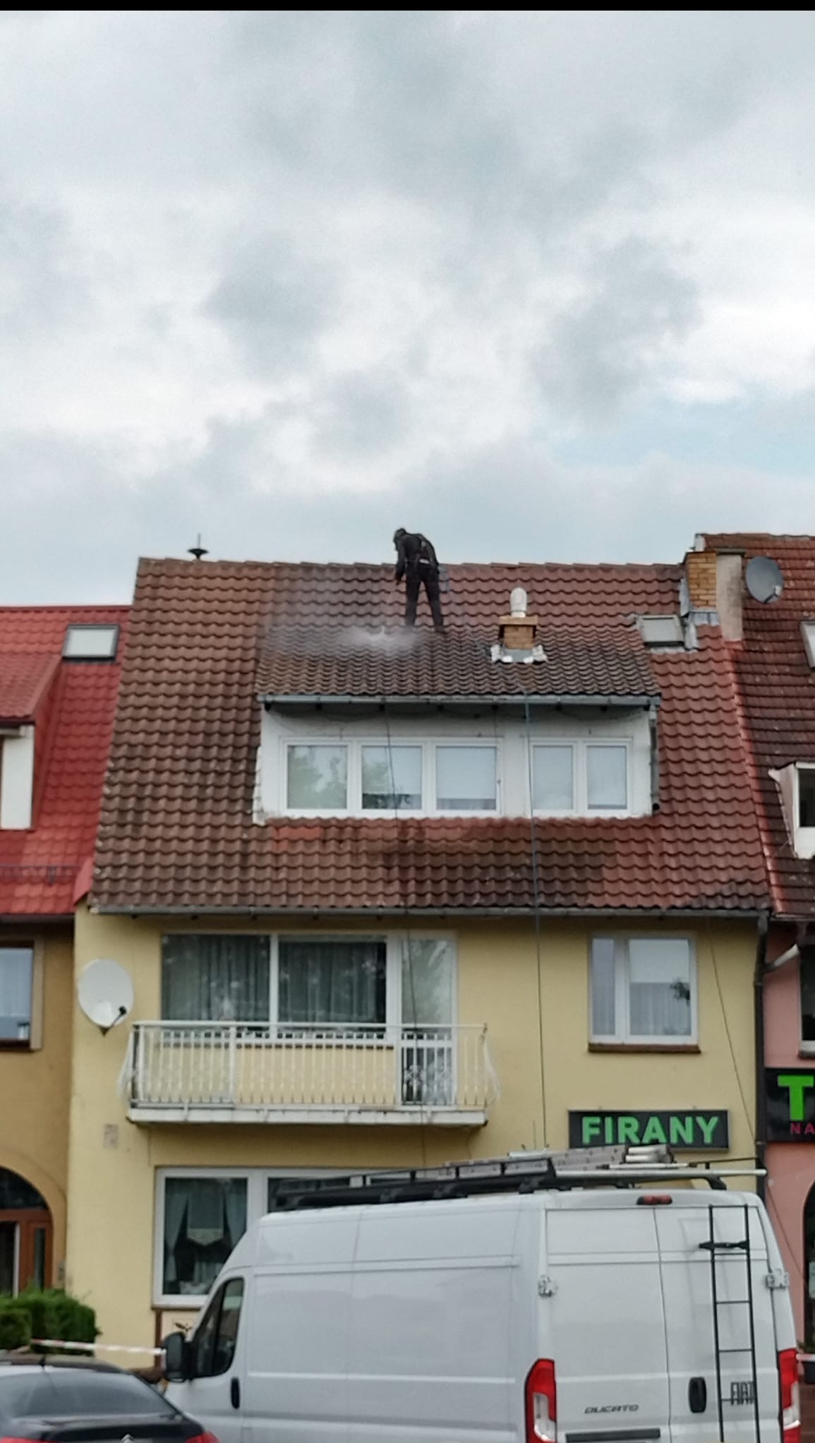 Pracownik w kombinezonie czyści dach budynku za pomocą myjki ciśnieniowej, unosząca się para wodna, widok z dołu na tle pochmurnego nieba.