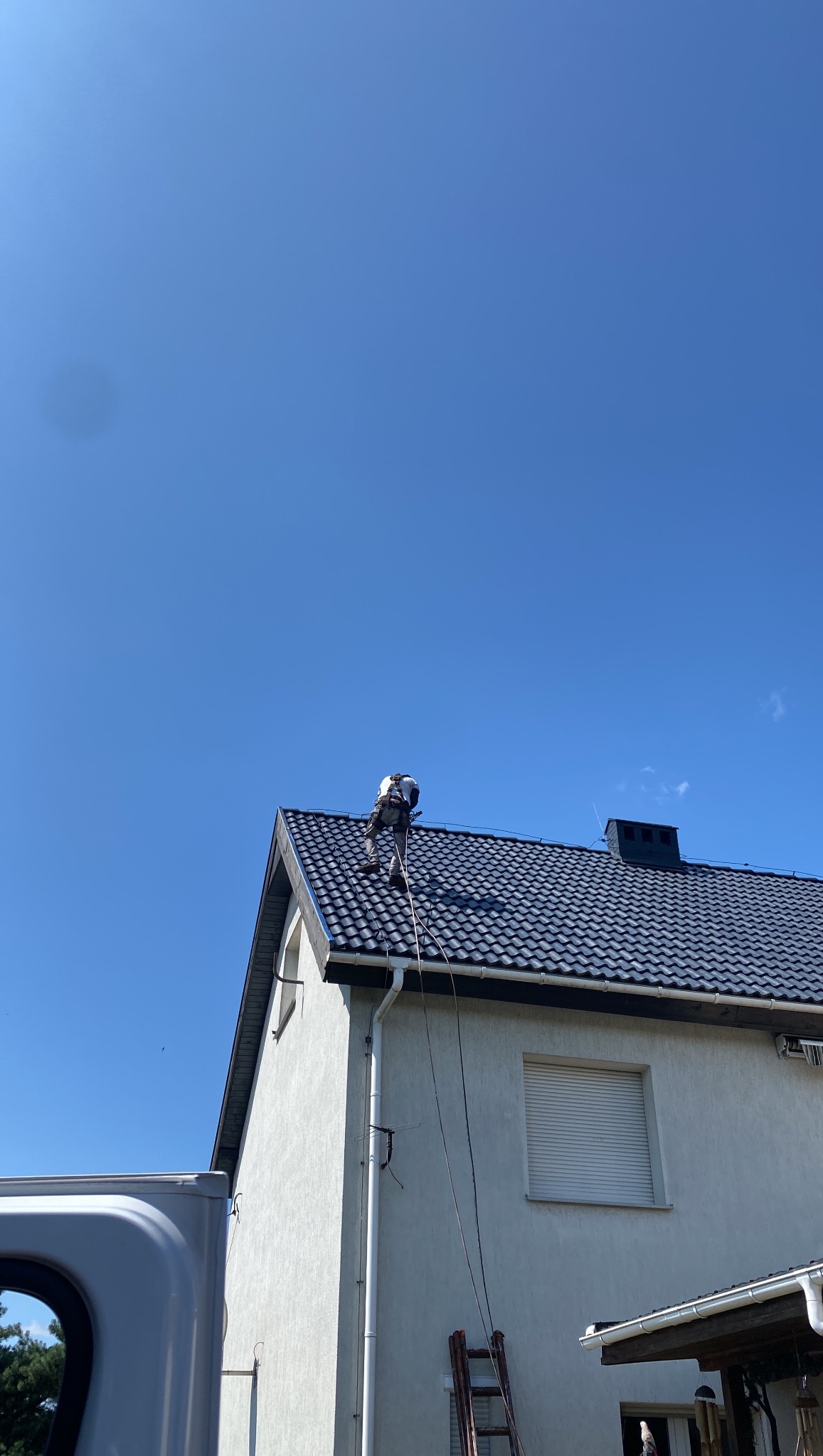 Pracownik w uprzęży na dachu domu z ciemną dachówką, pod błękitnym niebem. Widoczna drabina i fragment białej ściany budynku. Ujęcie z dołu, w słoneczny dzień.
