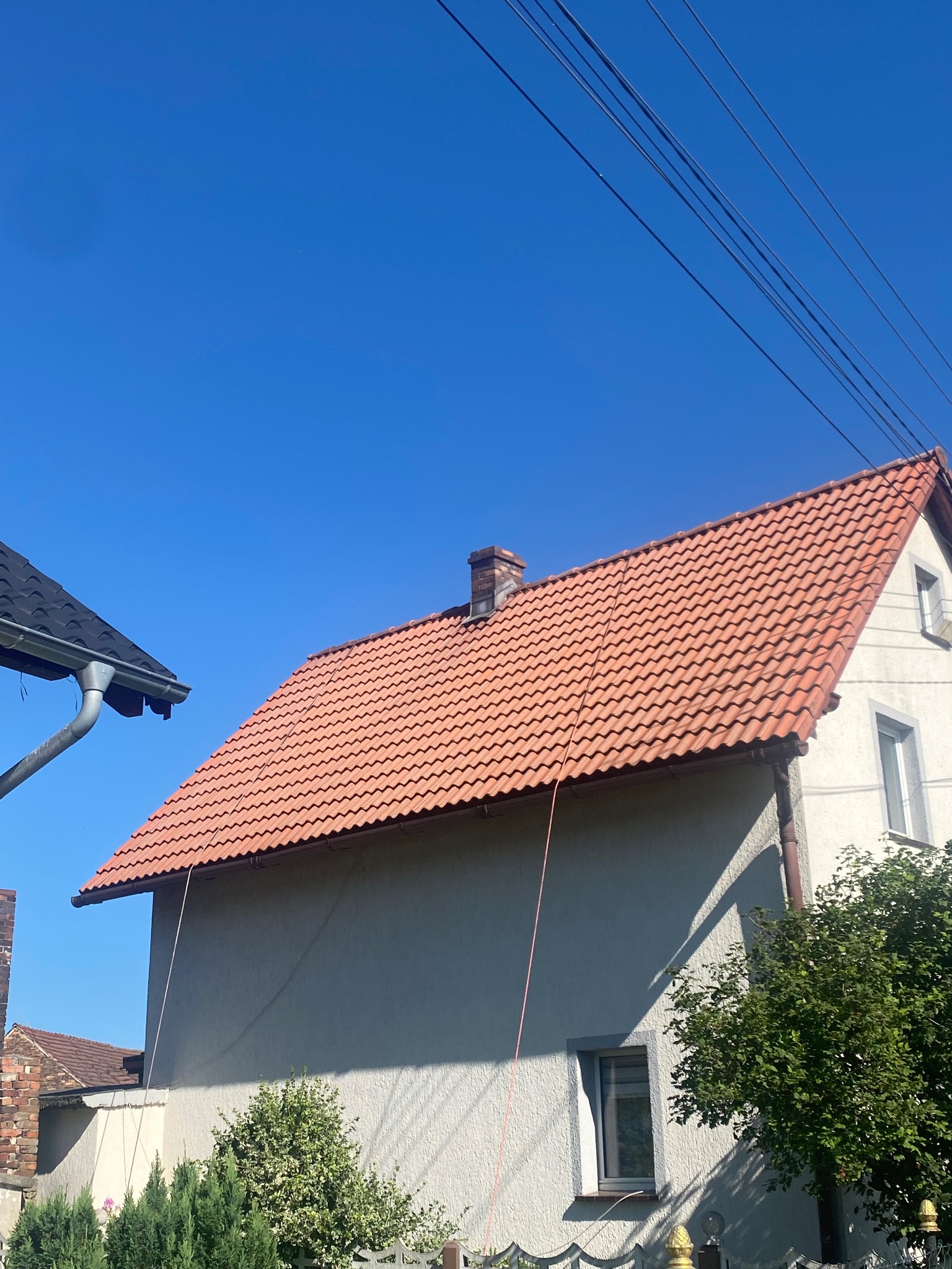 Dom jednorodzinny z czerwoną dachówką, widoczny z dołu w słoneczny dzień. Fragment budynku sąsiada i przewody elektryczne na tle błękitnego nieba.