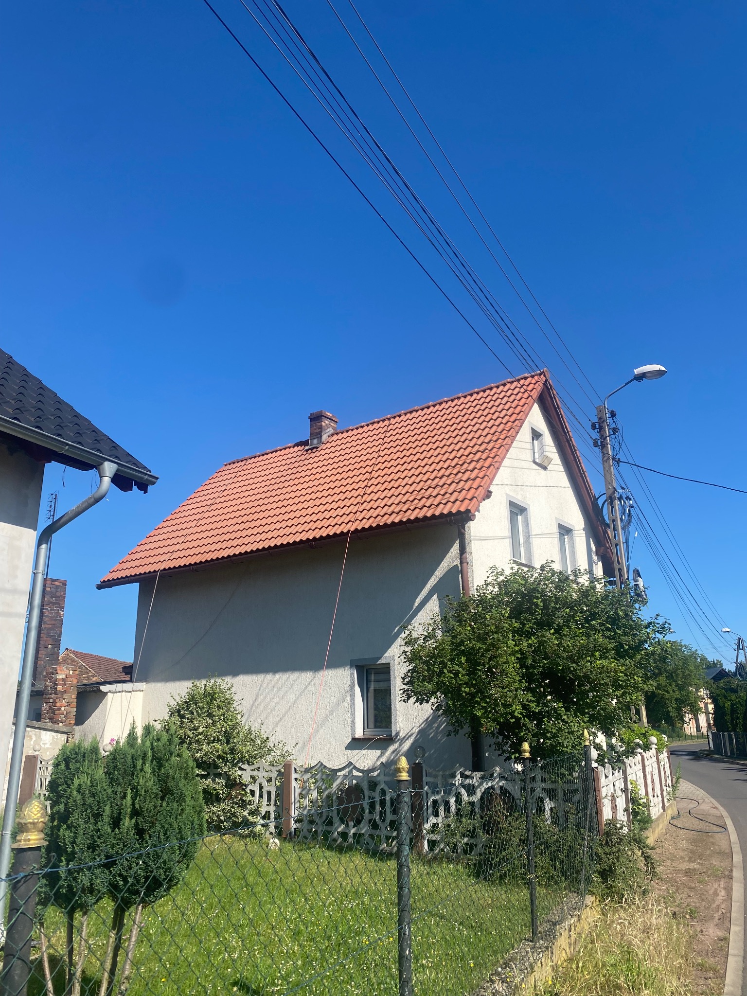 Dwupiętrowy dom z czerwonym dachem i jasną elewacją w słoneczny dzień. Widoczne okna, komin, drzewa i ogrodzenie. Na pierwszym planie trawa i ulica.