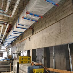 Arnor Budownictwo - Instalacja wentylacyjna w folii ochronnej, mocowana do sufitu. Widoczne materiały izolacyjne Isover i rusztowania. Stan surowy, cegła i beton.