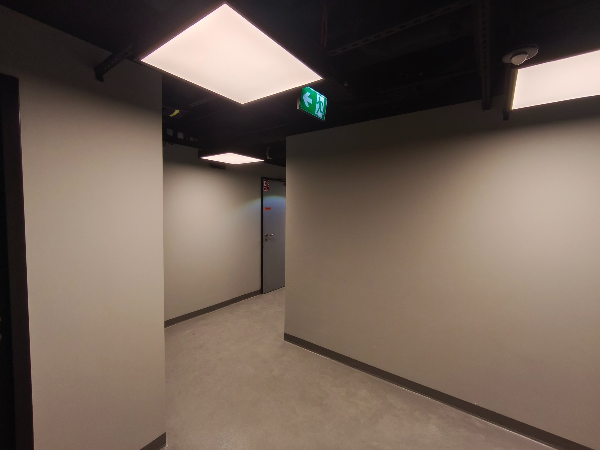 Korytarz biurowy z szarymi ścianami, betonową podłogą i kwadratowymi lampami sufitowymi. Widoczny znak ewakuacyjny.