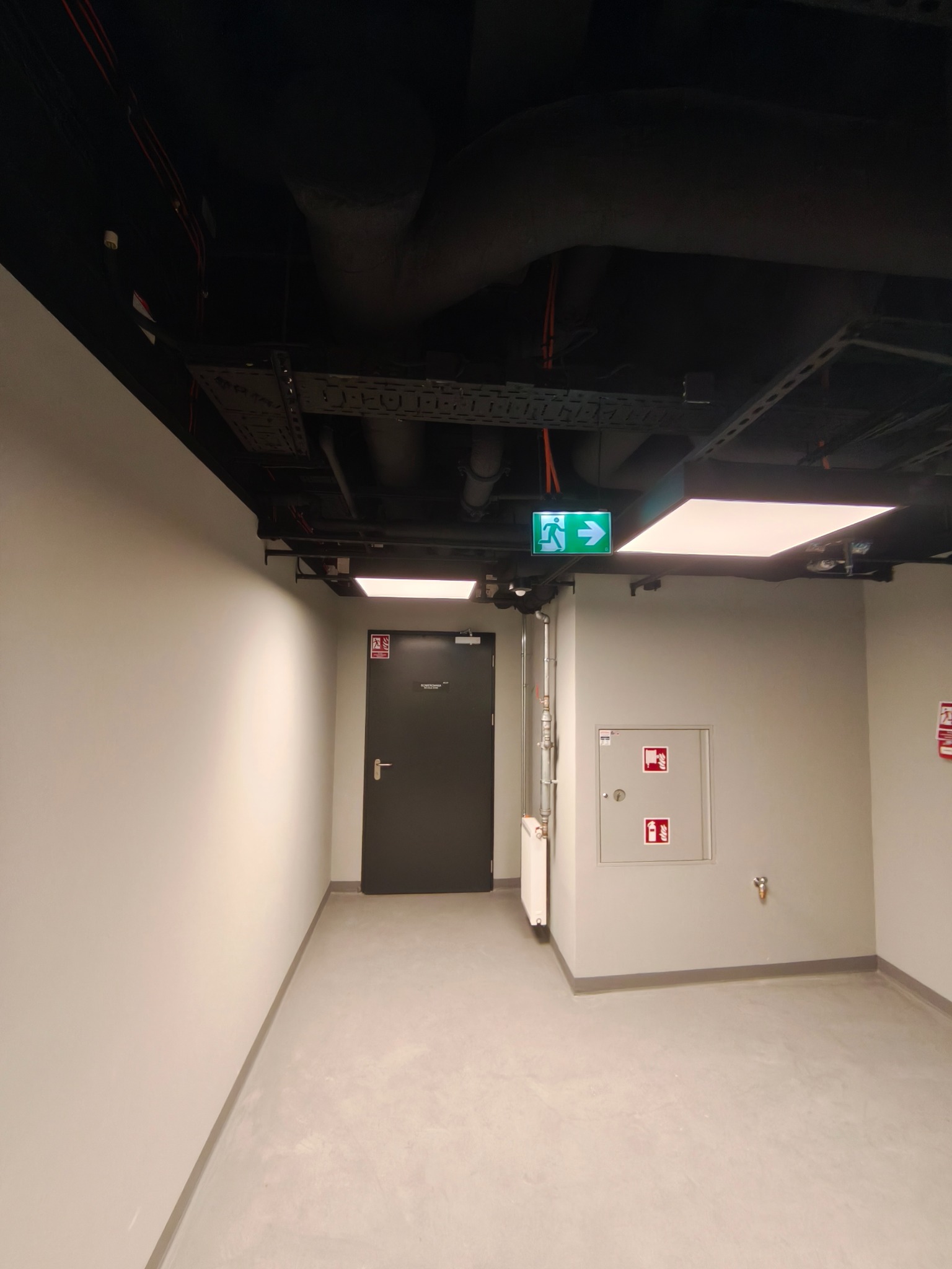 Korytarz biurowy z szarymi ścianami i podłogą, widoczne instalacje pod sufitem, znak ewakuacyjny, drzwi przeciwpożarowe i skrzynka hydrantowa.