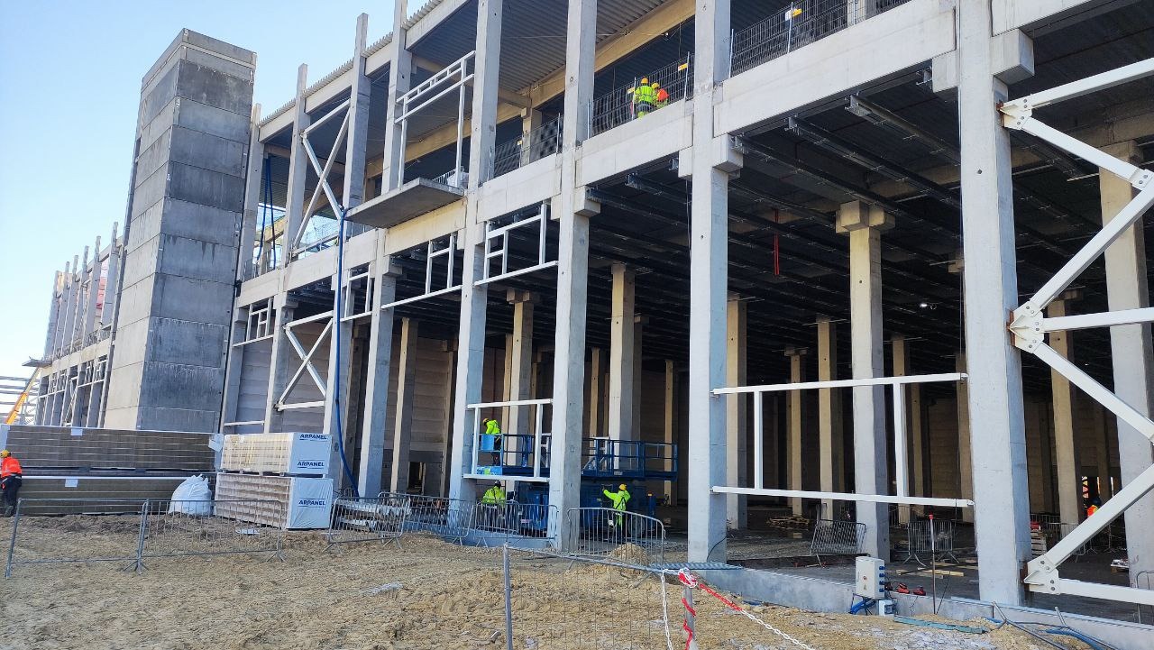 Wnętrze konstrukcji budynku w trakcie budowy. Widoczne betonowe filary, stalowe elementy konstrukcyjne, palety z materiałami izolacyjnymi i pracownicy w odblaskowych kamizelkach.