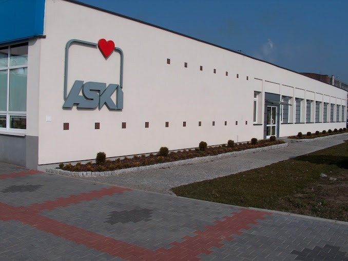 Fasada budynku firmy ASK z szarym logo zawierającym czerwone serce, rzędem okien i ozdobnymi krzewami wzdłuż ścieżki.