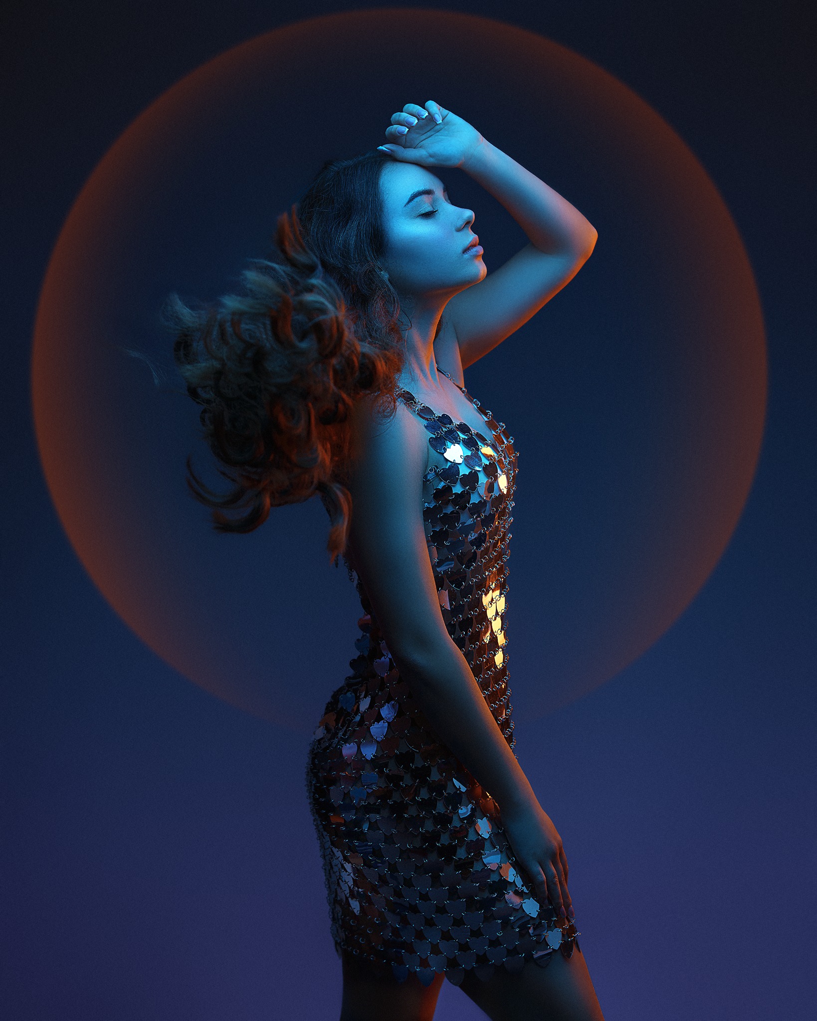Portret kobiety w srebrnej sukience na tle okręgu światła, z ręką na czole i zamkniętymi oczami. Ujęcie z profilu, oświetlenie w odcieniach niebieskiego i pomarańczowego.