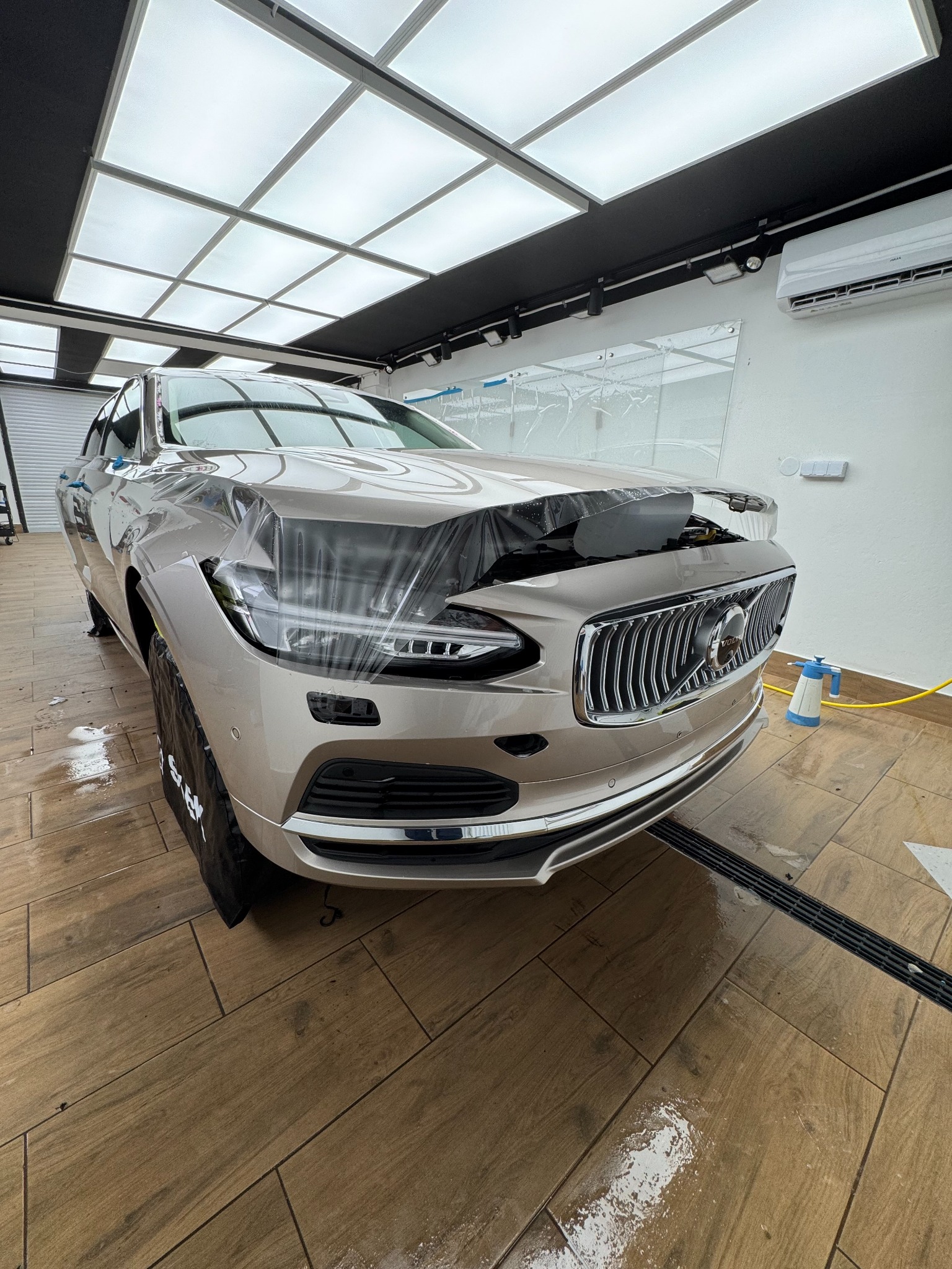 Jasny SUV Volvo w trakcie zabezpieczania folią ochronną w dobrze oświetlonym warsztacie. Widoczne detale grilla i reflektorów. Na podłodze płytki imitujące drewno.