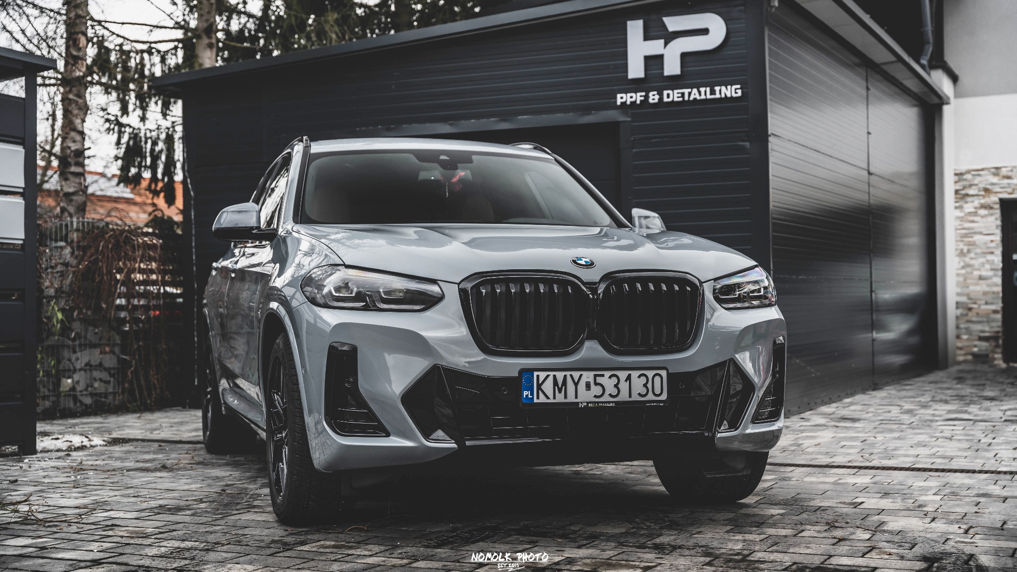 Szary BMW X3 po detailingu, zaparkowany przed garażem z logo 'HP PPF & Detailing'. Błyszcząca karoseria, czarny grill, polska tablica rejestracyjna. Profesjonalna fotografia.