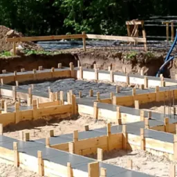 Wykopane fundamenty z drewnianym szalunkiem wypełnione betonem, widoczny pracownik w tle, zbrojenie, przygotowane pod dalsze etapy budowy.