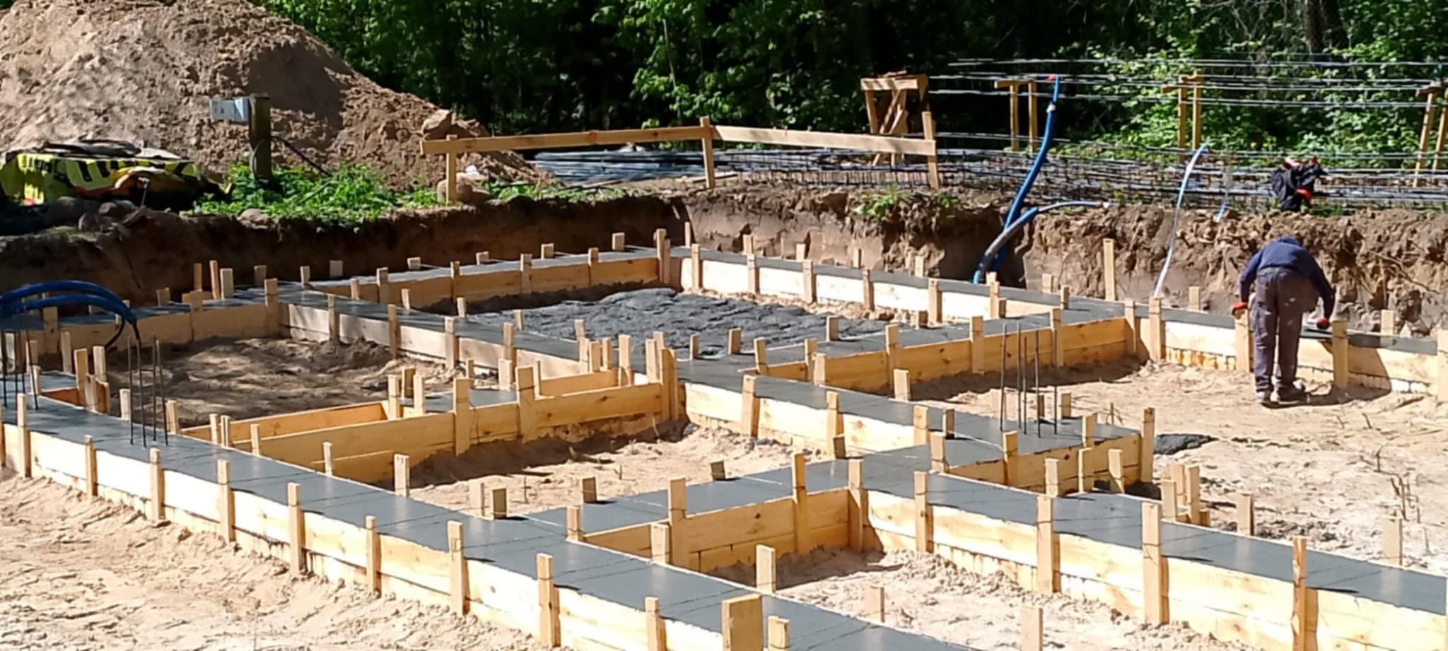 Wykopane fundamenty z drewnianym szalunkiem wypełnione betonem, widoczny pracownik w tle, zbrojenie, przygotowane pod dalsze etapy budowy.