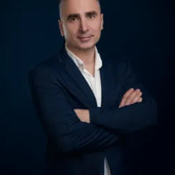 Portret agenta nieruchomości z Rogoża w granatowej marynarce, z założonymi rękami, na ciemnym tle. Profesjonalny wizerunek.