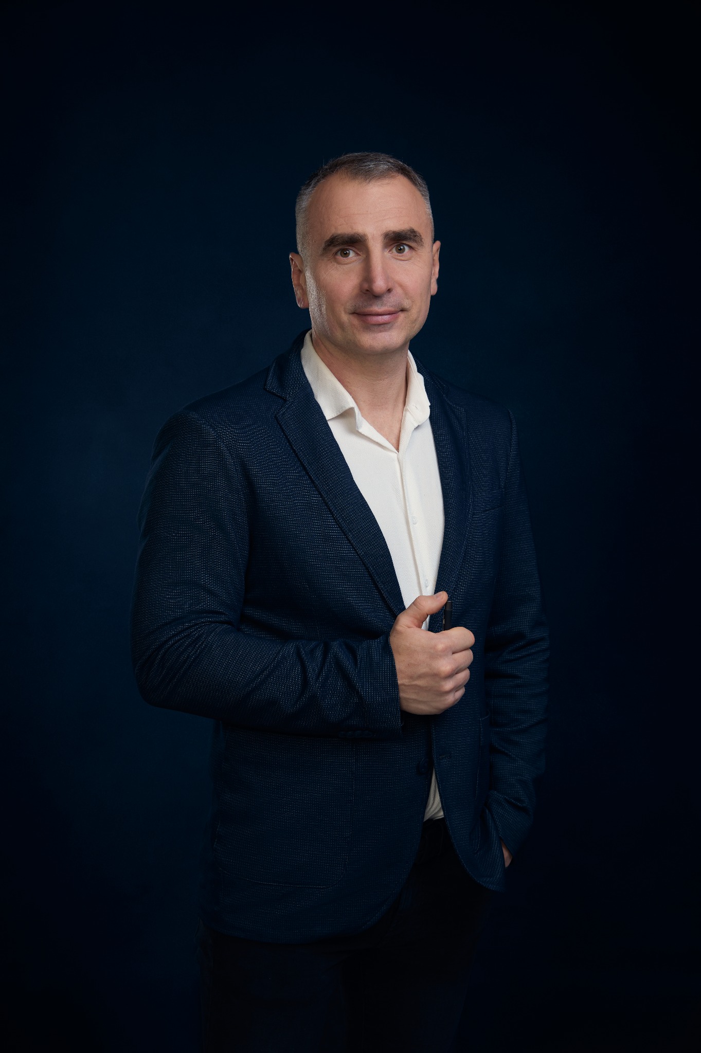 Portret biznesowy mężczyzny w granatowej marynarce i białej koszuli na ciemnym tle, z ręką w kieszeni i spokojnym uśmiechem.