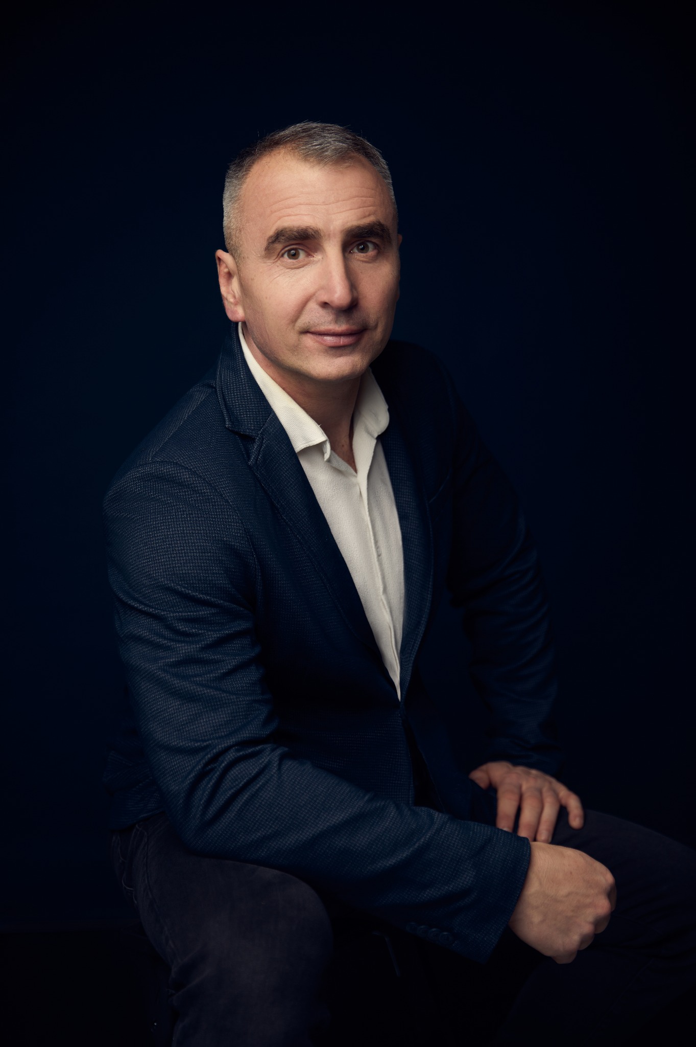 Portret mężczyzny w marynarce na ciemnym tle, siedzącego bokiem, z delikatnym uśmiechem, prezentujący profesjonalny wizerunek.