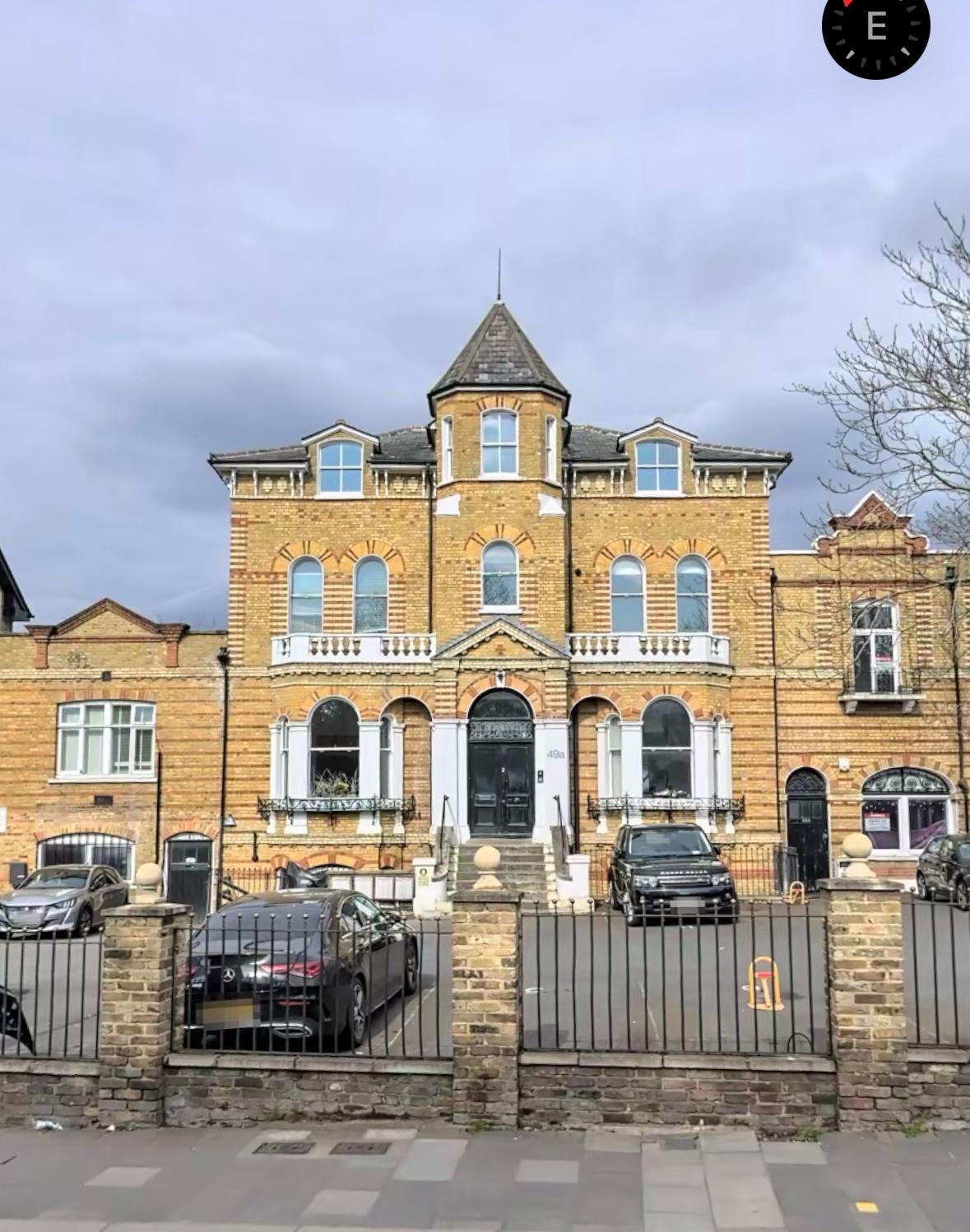 Żółta, ceglana kamienica z wieżyczką, balkonami i czarnymi drzwiami wejściowymi. Przed budynkiem zaparkowane samochody i ogrodzenie z cegły i metalu.