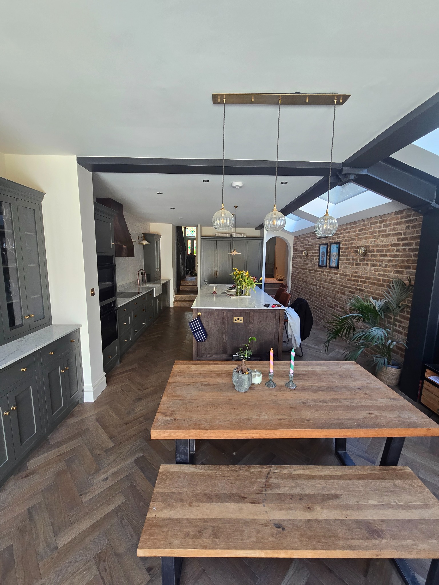 Stylowe wykończenie wnętrza kuchni z wyspą, drewnianym stołem i parkietem ułożonym w jodełkę. Widoczny fragment przeszklonego dachu i ceglanej ściany.