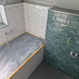 Wnętrze łazienki w trakcie prac wykończeniowych. Widoczne płytki w odcieniach bieli i zieleni, zabezpieczona folią wanna i fragment podłogi. Surowy stan ścian.