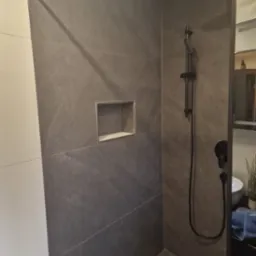 Nowoczesna kabina prysznicowa z szarymi płytkami imitującymi kamień, czarną armaturą i wnęką w ścianie, ukazująca minimalistyczny design wykończenia łazienki.