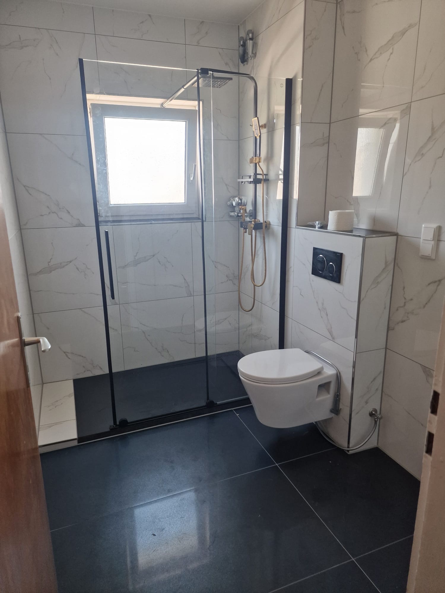 Nowoczesna łazienka z kabiną prysznicową typu walk-in, toaletą podwieszaną i marmurowymi płytkami. Czarna armatura i ciemna podłoga kontrastują z jasnymi ścianami.