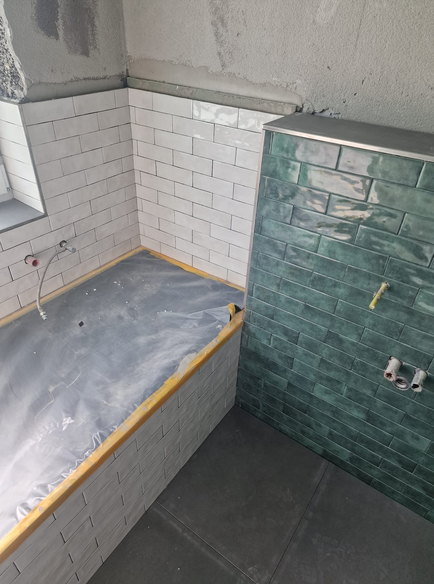 Wnętrze łazienki w trakcie prac wykończeniowych. Widoczne płytki w odcieniach bieli i zieleni, zabezpieczona folią wanna i fragment podłogi. Surowy stan ścian.