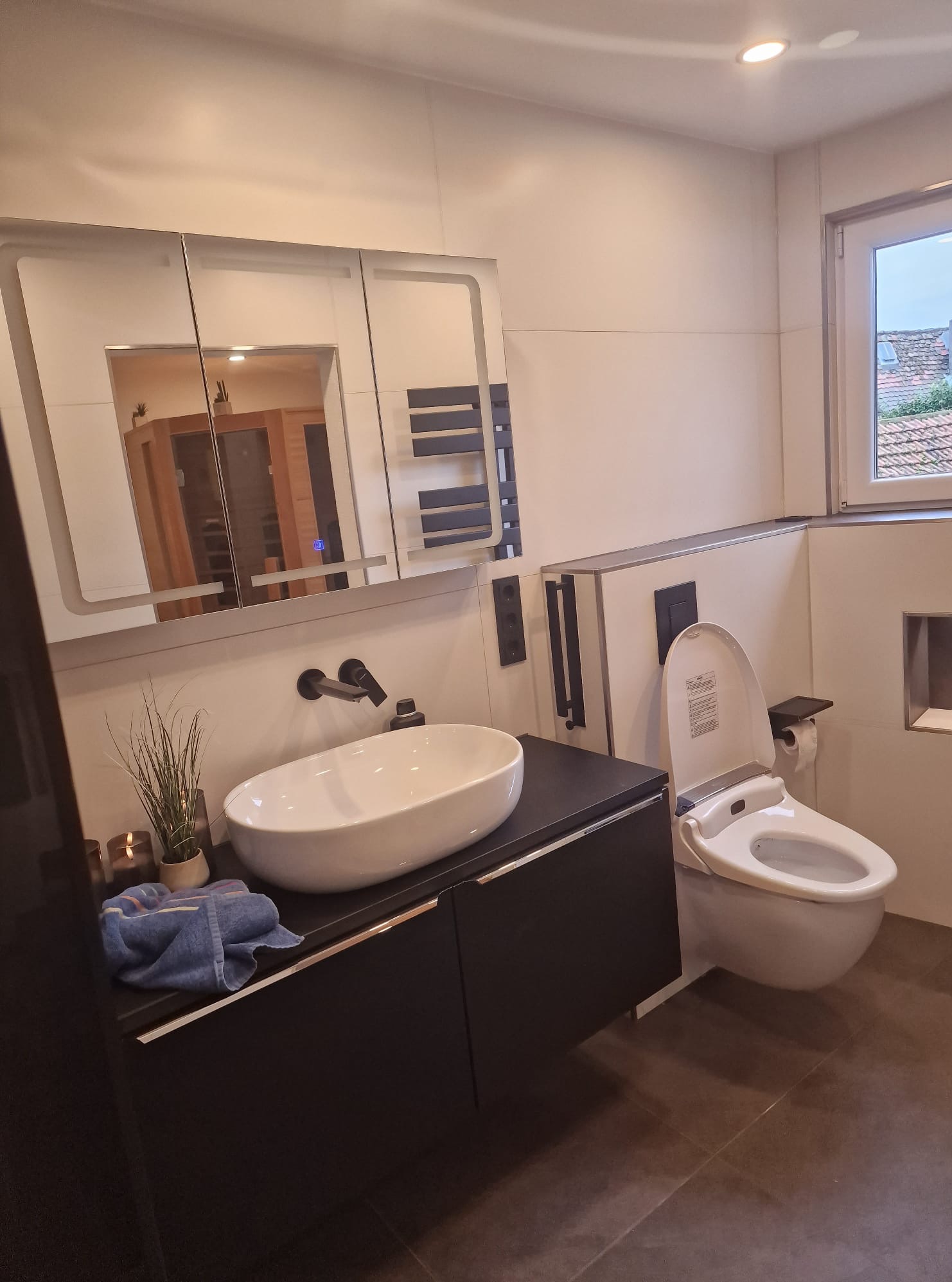 Nowoczesna łazienka z wiszącą toaletą z funkcją bidetu, umywalką nablatową na czarnej szafce i lustrem z podświetleniem LED. Widoczne płytki podłogowe w odcieniach brązu.