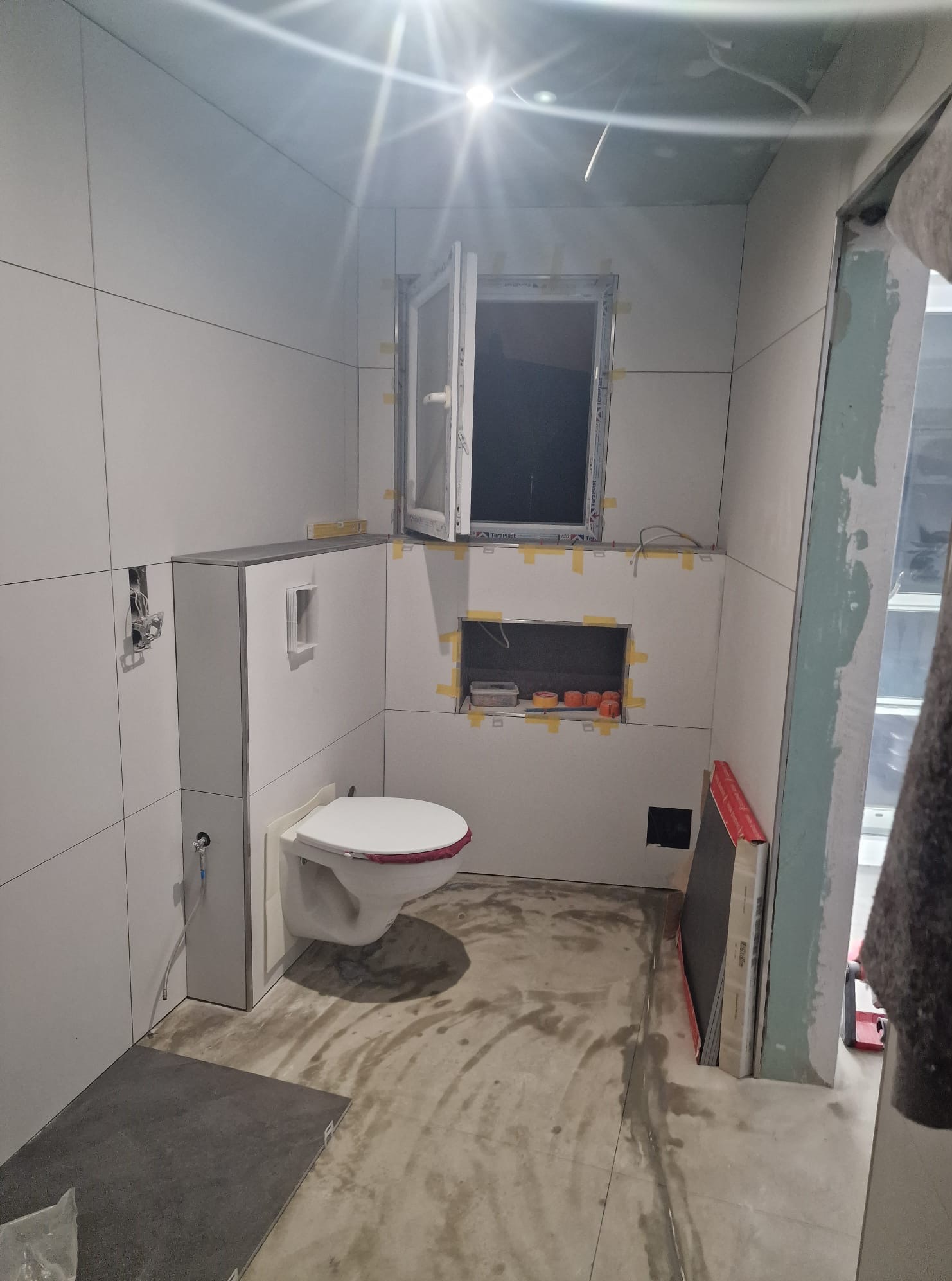 Biała, częściowo wykończona łazienka z podwieszaną toaletą, oknem i wnęką z pomarańczowymi elementami. Widoczne płytki i przygotowane podłoże pod podłogę.