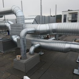 ICE VENTURO SPÓŁKA Z OGRANICZONĄ ODPOWIEDZIALNOŚCIĄ - Skomplikowany system srebrnych, metalowych rur wentylacyjnych na dachu budynku, z widocznymi elementami montażowymi i fragmentem budynku w tle.