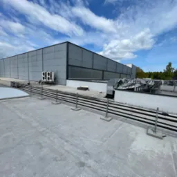 Widok z góry na płaski dach budynku komercyjnego z instalacją wentylacyjną, klimatyzatorami i elementami zabezpieczającymi. Błękitne niebo z chmurami w tle.