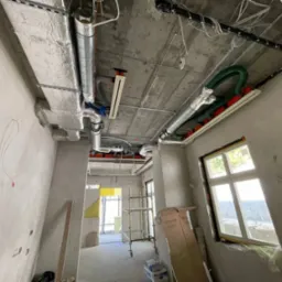 Wnętrze w trakcie montażu instalacji wentylacyjnej: widoczne rury wentylacyjne pod sufitem, surowy beton, ściany w tynku i okno z widokiem na zewnątrz.