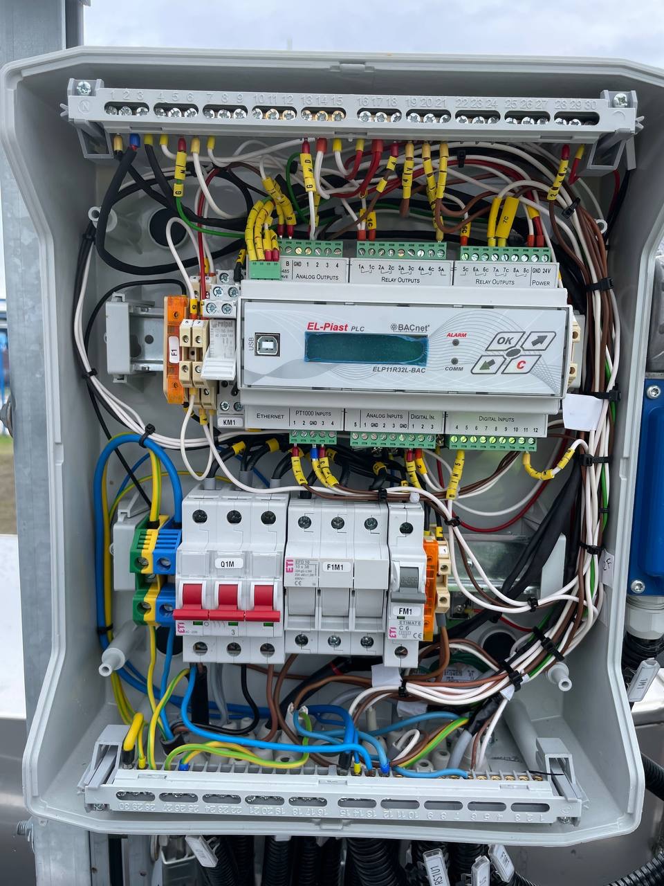 Otwarta szafka sterownicza z widocznymi przewodami, bezpiecznikami i modułem PLC El-Plast PLC ELP11R32L-BAC. Widoczne złącza, kable w kolorach żółtym, niebieskim i brązowym.