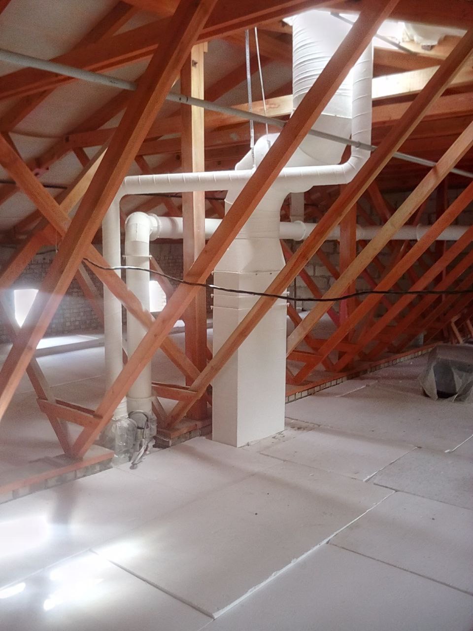 Białe rury rekuperacji w surowym wnętrzu poddasza z drewnianą konstrukcją więźby dachowej. Elementy instalacji w pionie i poziomie, widoczne łączenia.