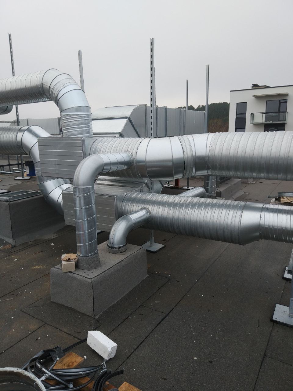 Skomplikowany system srebrnych, metalowych rur wentylacyjnych na dachu budynku, z widocznymi elementami montażowymi i fragmentem budynku w tle.