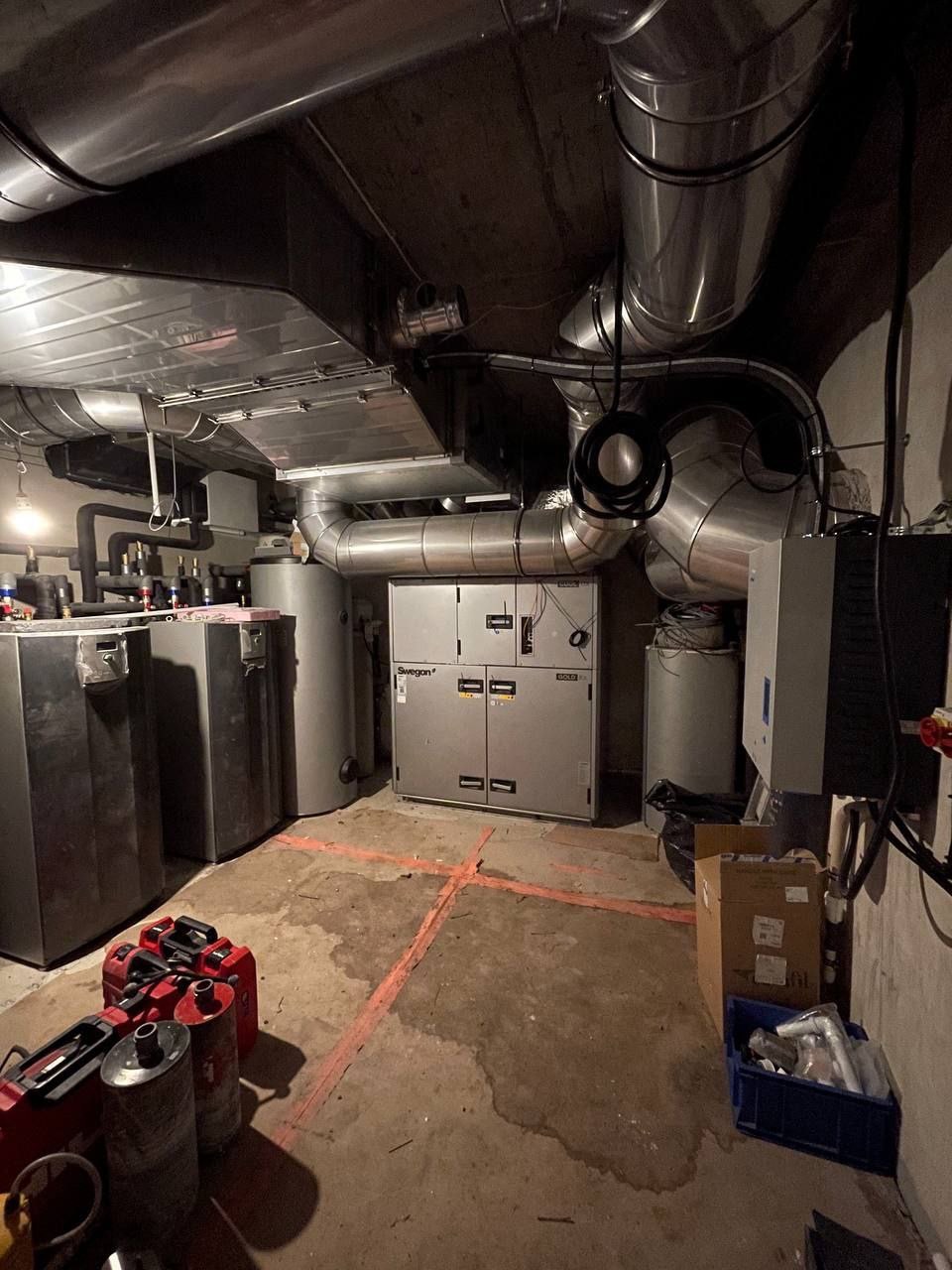 Piwnica z centralą wentylacyjną Swegon, rurami wentylacyjnymi pod sufitem i narzędziami na podłodze. Widoczne zbiorniki i skrzynki z akcesoriami.