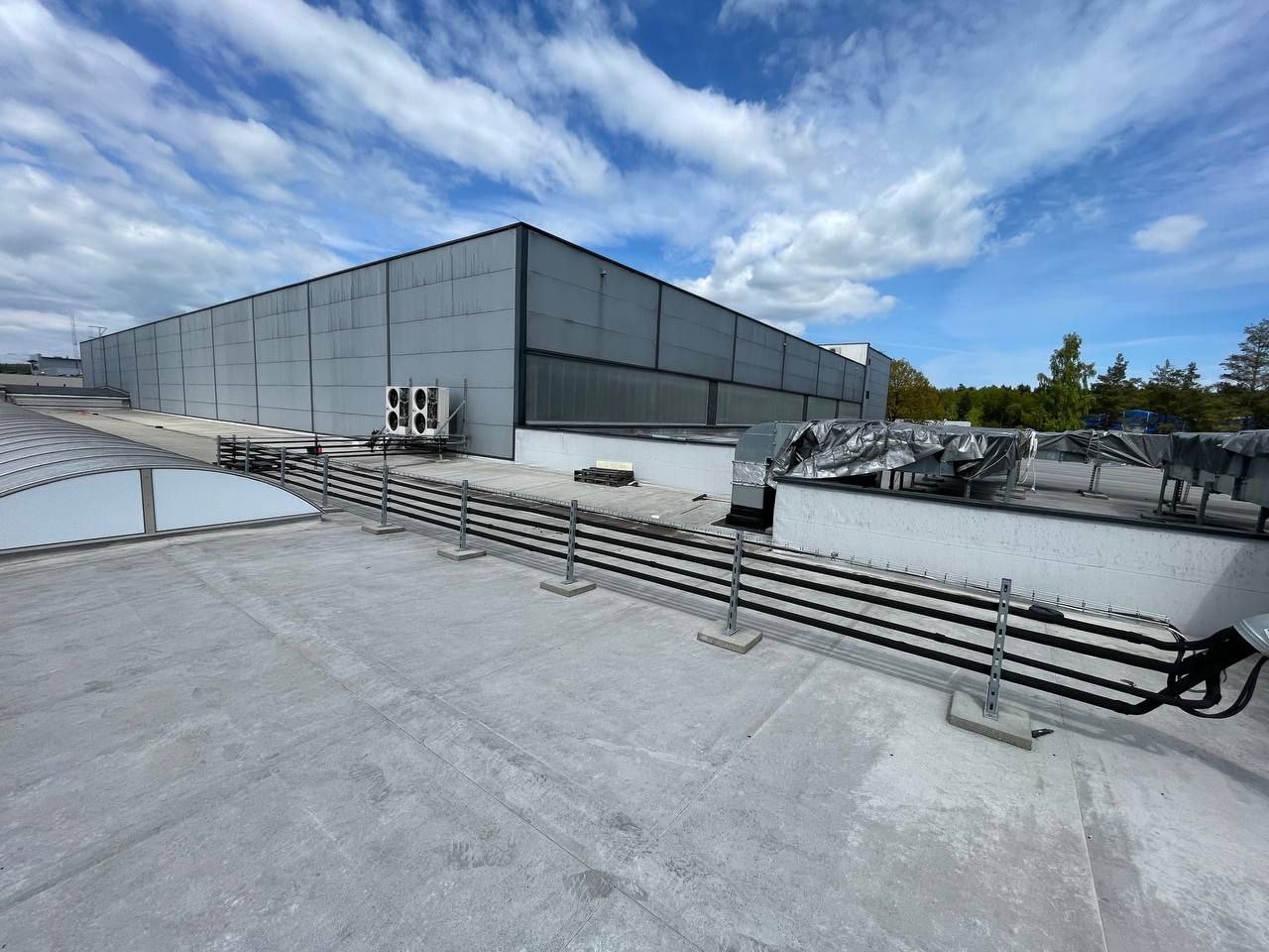 Widok z góry na płaski dach budynku komercyjnego z instalacją wentylacyjną, klimatyzatorami i elementami zabezpieczającymi. Błękitne niebo z chmurami w tle.