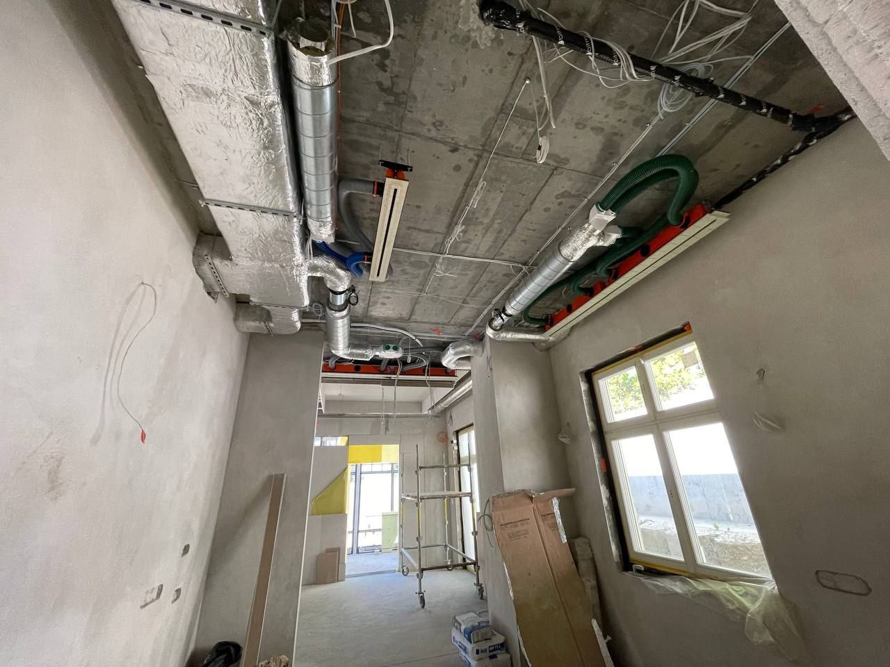 Wnętrze w trakcie montażu instalacji wentylacyjnej: widoczne rury wentylacyjne pod sufitem, surowy beton, ściany w tynku i okno z widokiem na zewnątrz.