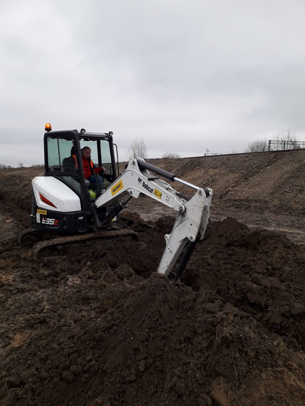 Operator w minikoparce Bobcat E35Z kopie dół w ziemi. W tle widoczne są tory kolejowe i pochmurne niebo. Praca w toku, teren przygotowany pod instalacje.